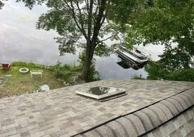 A roof with a skylight overlooks a lake with a pontoon boat. Green trees and grass line the shore.