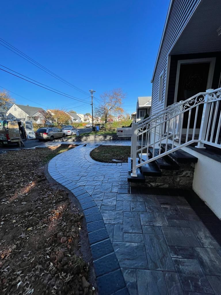 A newly paved walkway curves toward a home's steps, featuring dark gray stones and a white railing under a blue sky.