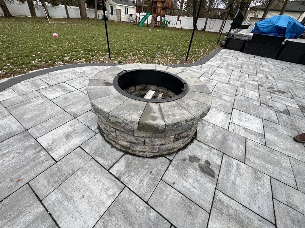 A stone fire pit sits on a paved patio in a backyard. The pit is surrounded by gray pavers.