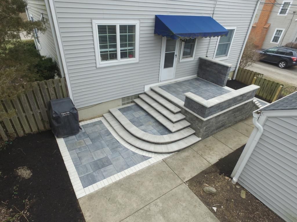 Gray house exterior with a newly constructed patio and steps, featuring gray pavers, a blue awning, and a retaining wall.