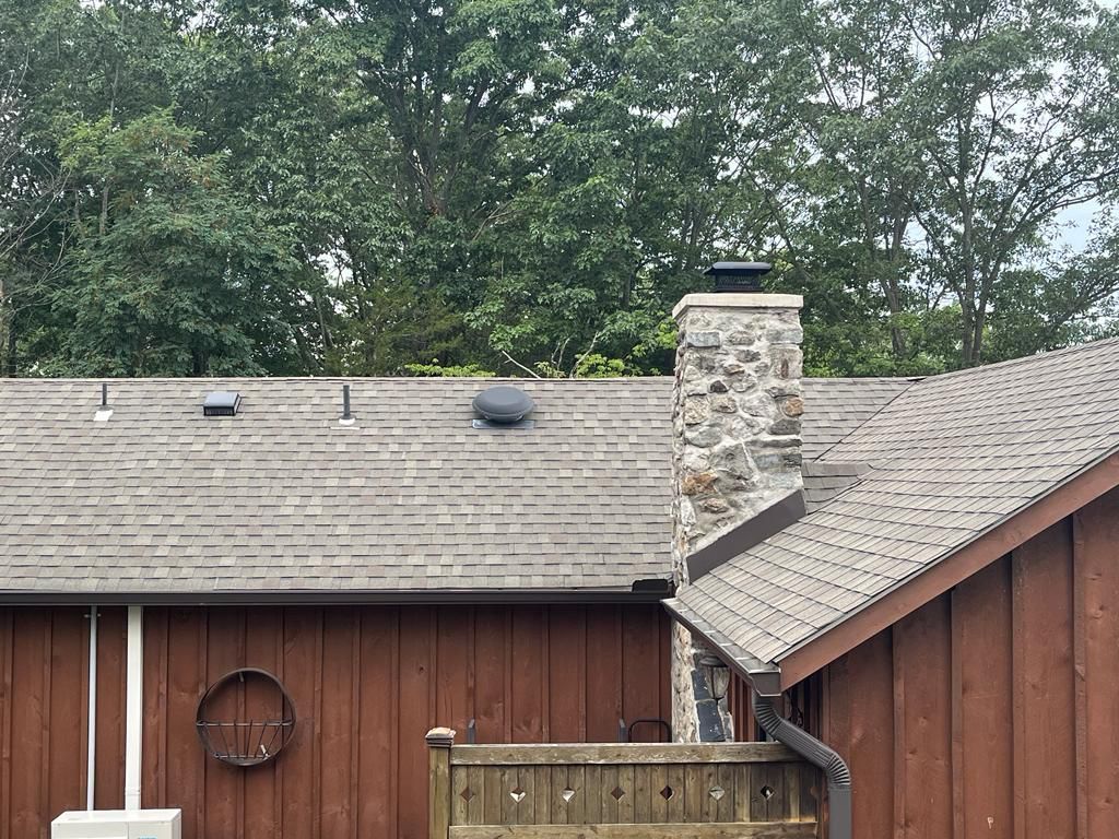 Exterior view of a brown building with a stone chimney, gray roof, and trees in the background.