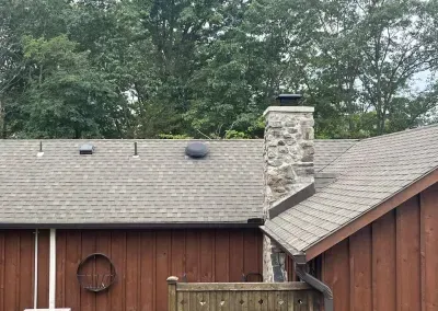 Brown wooden building with a stone chimney on a gray shingle roof. Green trees are in the background.