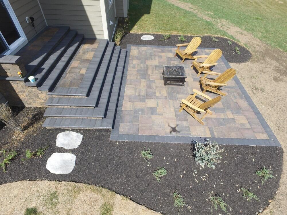A paved patio with steps leading from a house, featuring Adirondack chairs, a fire pit, and landscaping.