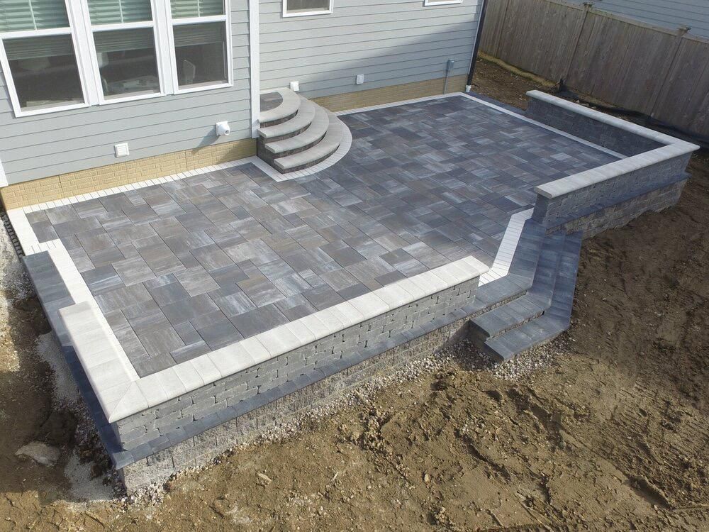 A newly constructed outdoor patio with gray pavers, retaining walls, and steps. The patio sits next to a light gray house.
