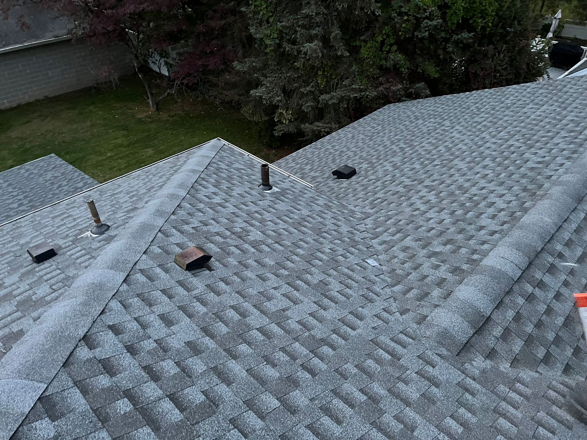 View of a gray shingled roof with several vents and a glimpse of greenery in the background.