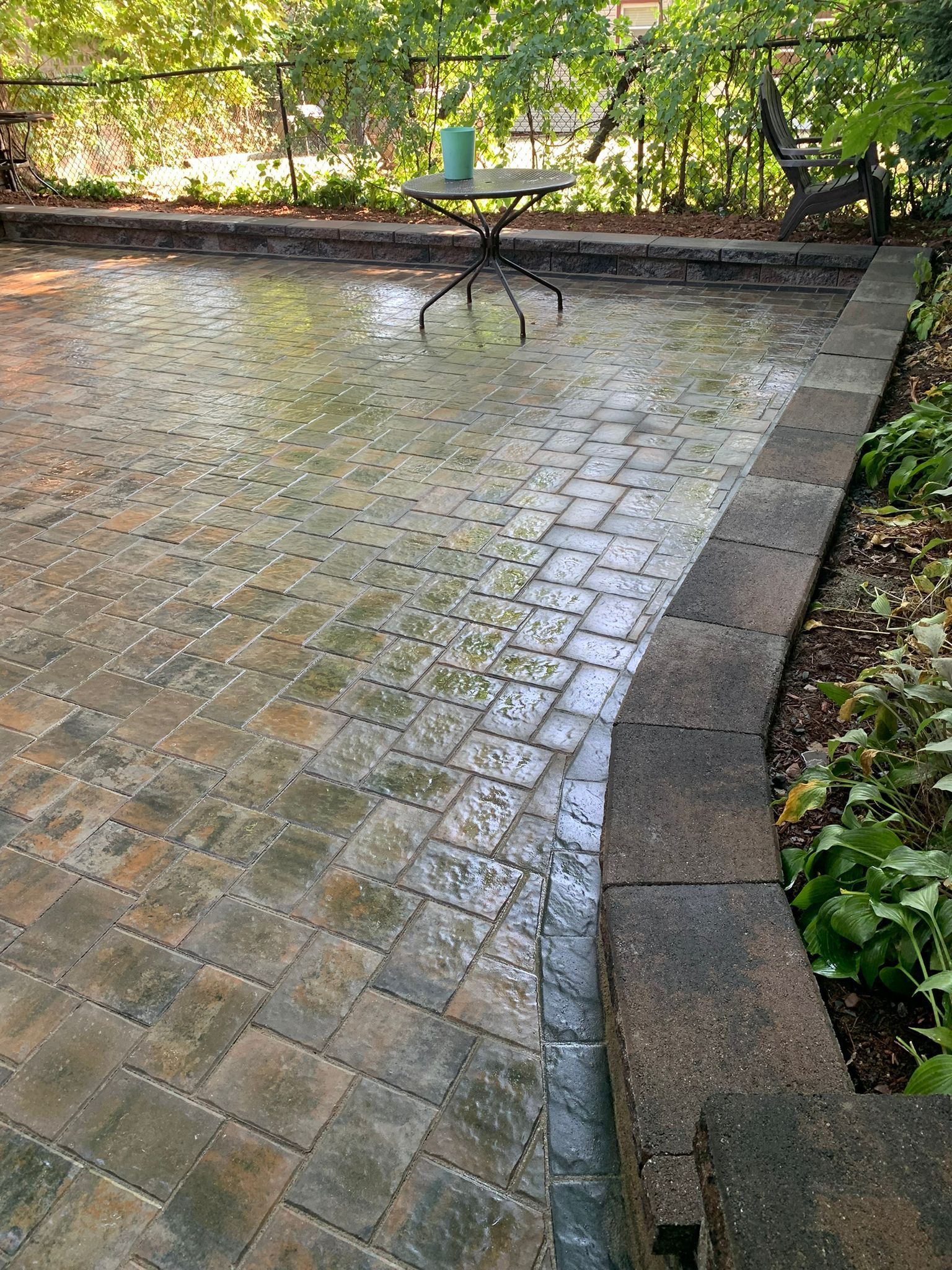 Rain-covered brick patio with a small table and surrounding greenery. A low stone wall borders one side of the patio.