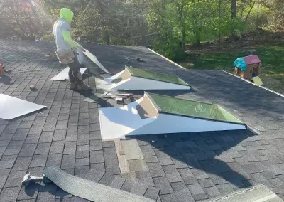 A roofer in green protective gear working on skylights on a shingled roof, sunny outdoor setting.