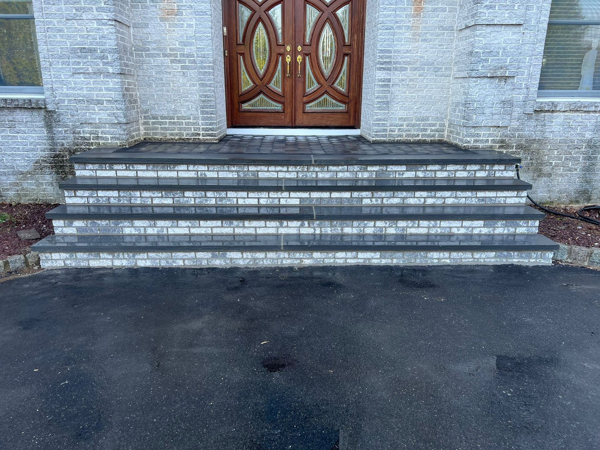 Three stone steps leading to a wooden double door. The steps have a gray and white brick pattern, and the asphalt driveway is wet.