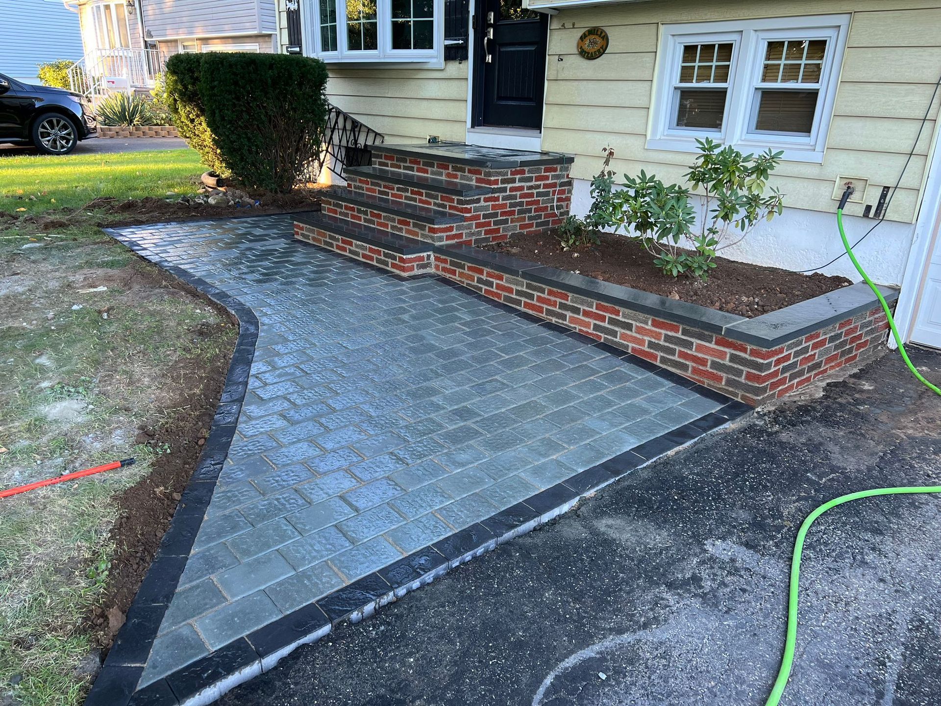 A brick and paver walkway and steps leading to a yellow house's front door. A dark car is parked to the left.