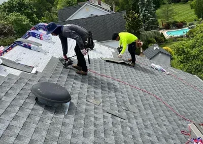 Two roofers working on a gray shingle roof. One kneels, the other stands, both are removing shingles. Green trees and a swimming pool are visible in the background.