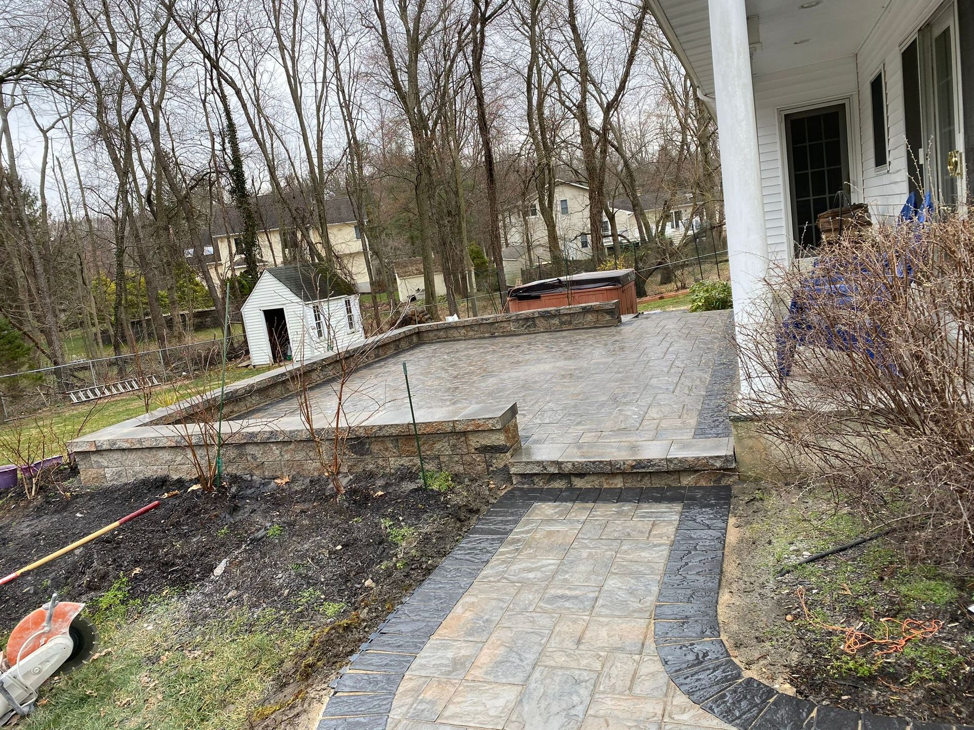 A brick patio and walkway in front of a white house, with a small shed and trees in the background.