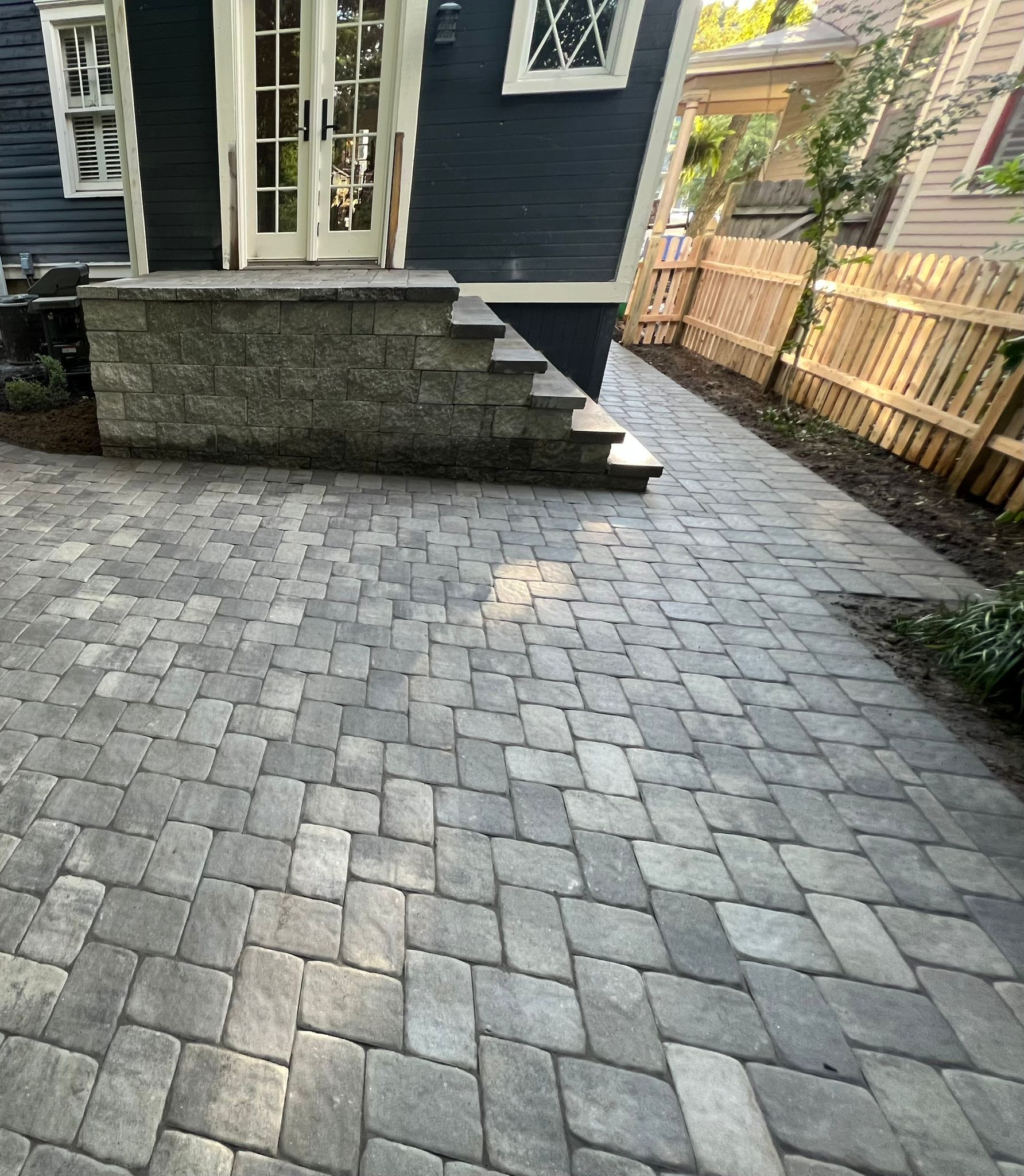 A paved patio with a dark blue house and stone steps. A wooden fence and walkway are to the right.