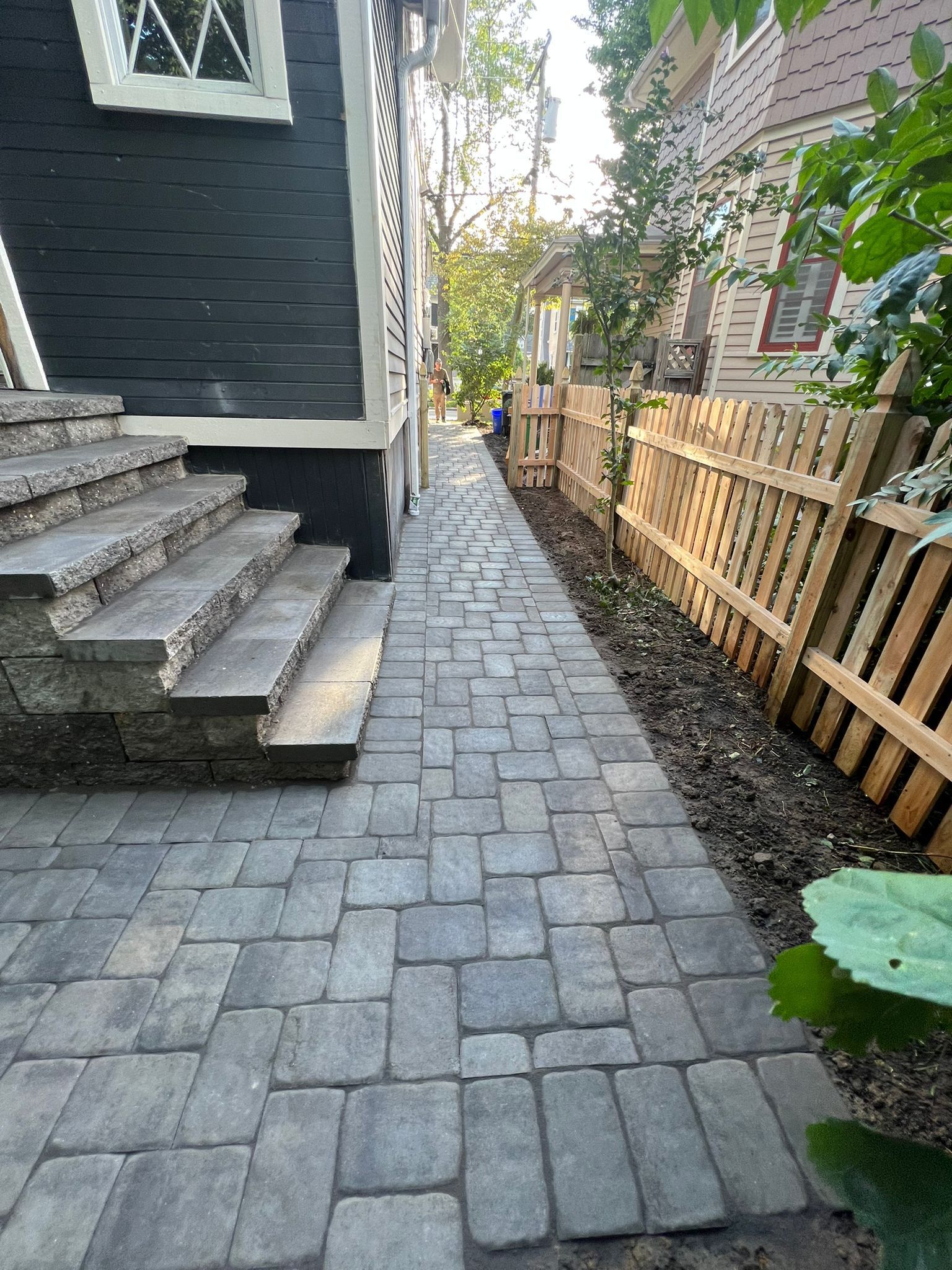 Brick walkway along a house with steps leading to a door. Wooden fence on the right side.