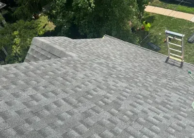 Gray shingle roof with a ladder and a worker in the background on a sunny day.
