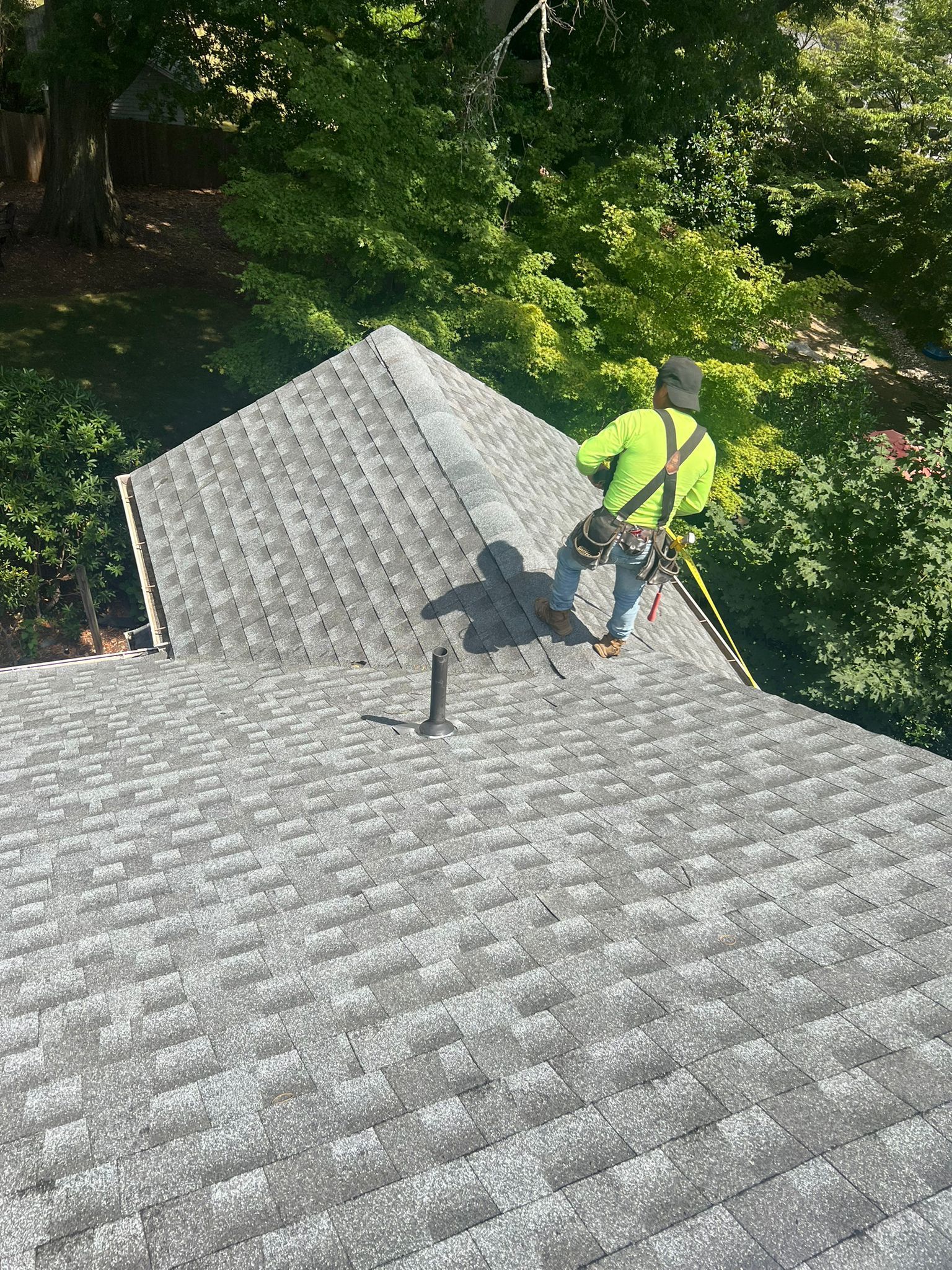 A worker on a gray shingled roof wears safety gear. He's against a backdrop of green trees.