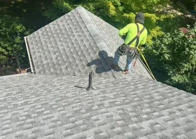 Roofer on a gray shingle roof wearing safety harness, with green safety vest. Trees in the background.