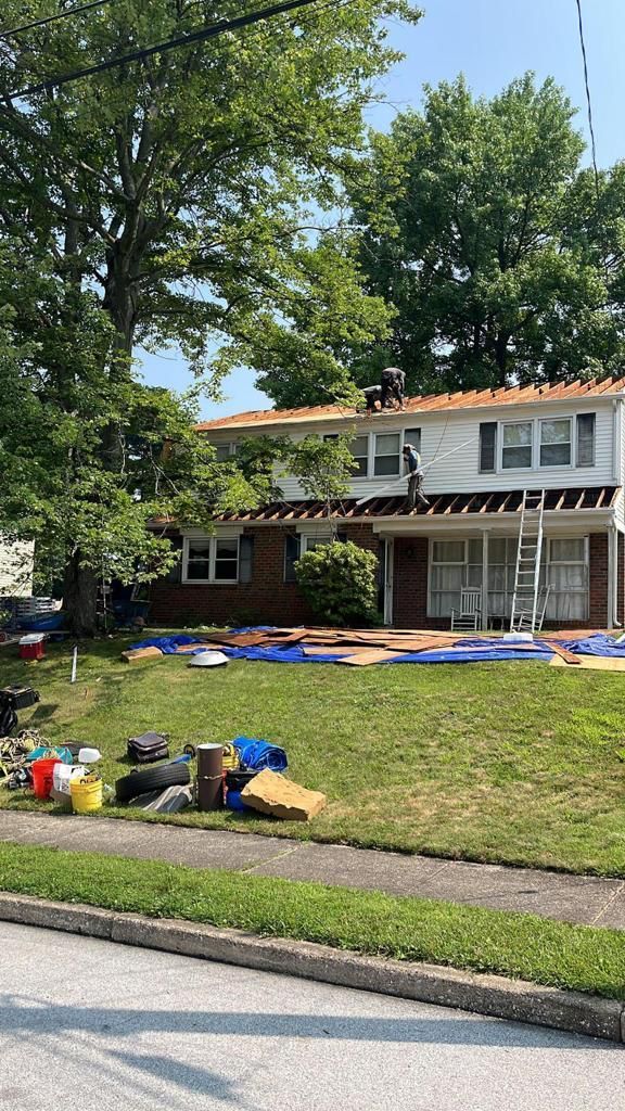 Two-story house under roof repair. Workers on the roof, covered with partially installed orange roofing tiles. Blue tarps and equipment are on the lawn.