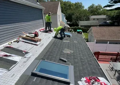 Roofers in safety vests working on a rooftop with shingles, tools, and a skylight on a sunny day.