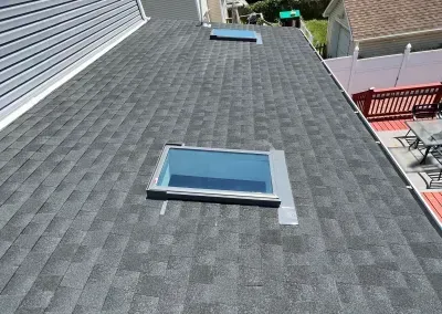 Gray asphalt shingle roof with two skylights, next to light blue siding and a deck with a red railing.