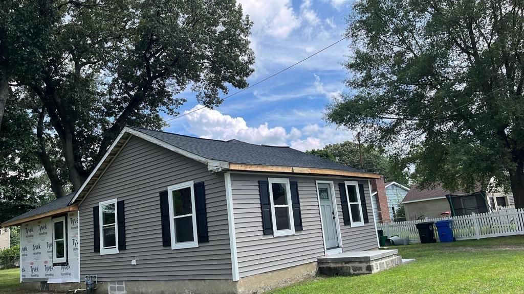Gray house with new roof and siding, black shutters, and a small stone step leading to the front door. Trees and blue sky in the background.