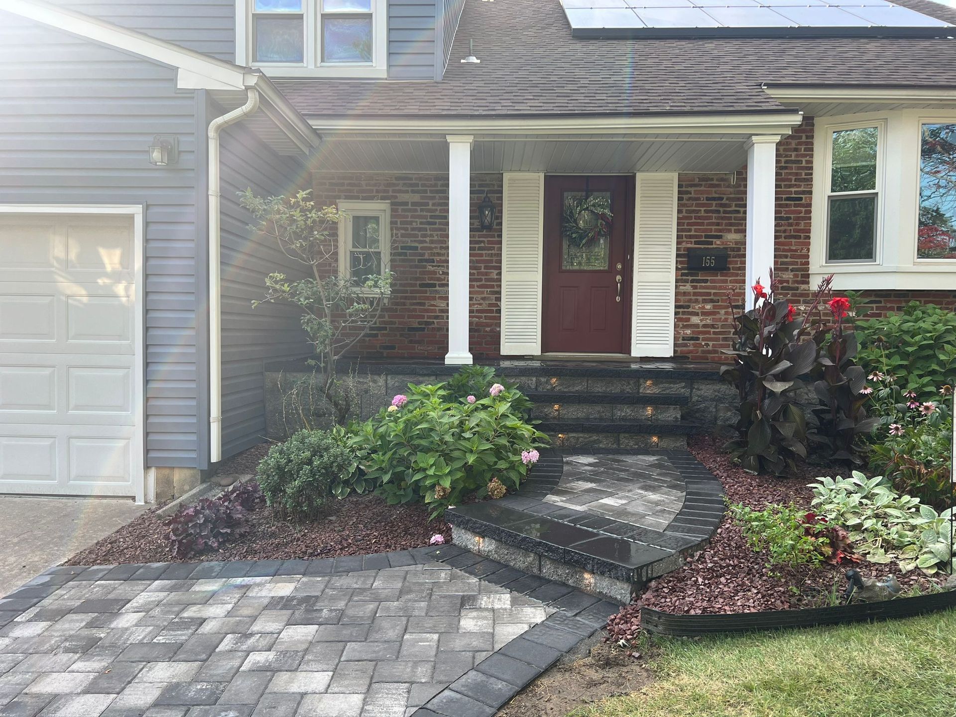 A house with a brick walkway and steps leading to a burgundy door. Landscaping includes various plants and flowers.