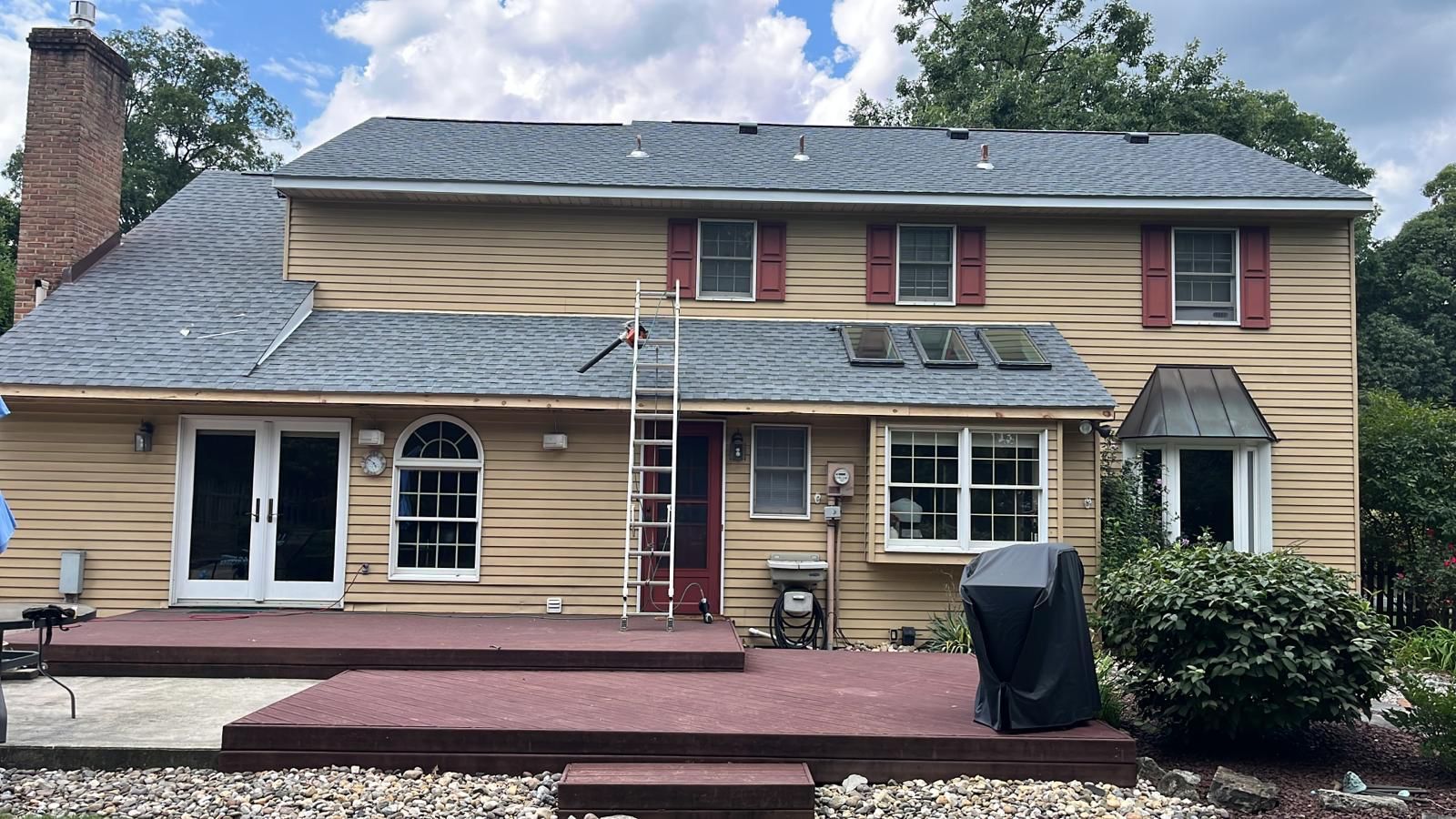 Backyard view of a two-story house with new gray roof, wooden siding, red shutters, and a wooden deck. A ladder is leaning against the house.