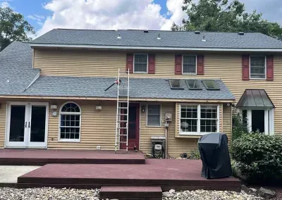 Back of a two-story tan house with a dark gray roof, red shutters, and a wooden deck. A ladder leans against the house.