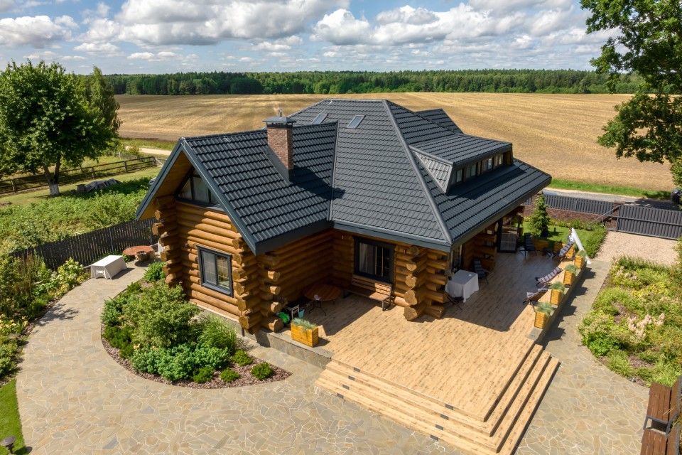 An aerial view of a rustic log cabin with a dark shingled roof, set on a stone patio surrounded by fields and greenery.
