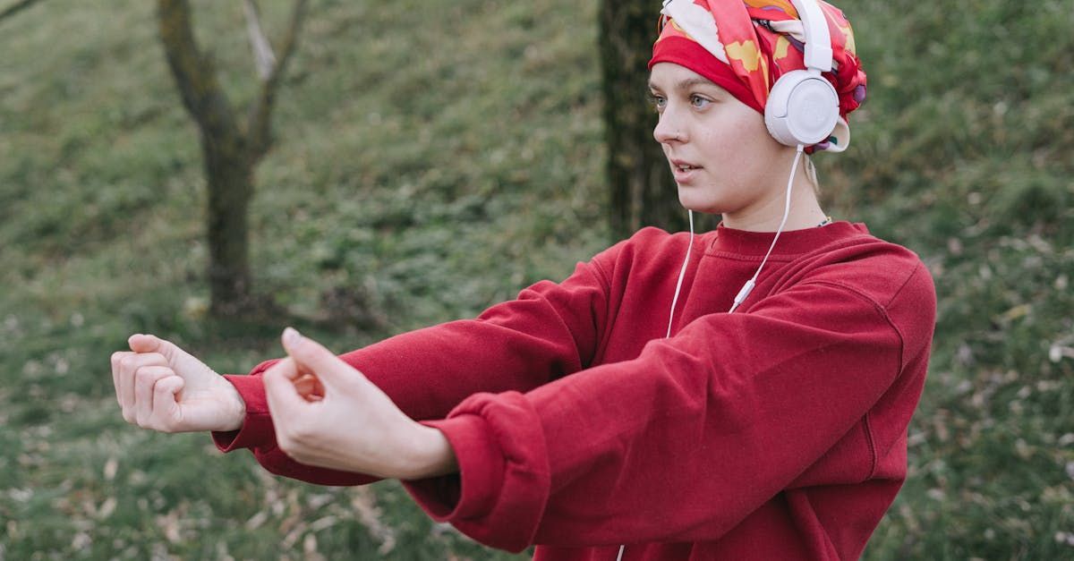 A woman wearing headphones and a red sweater is stretching her arms in the woods.