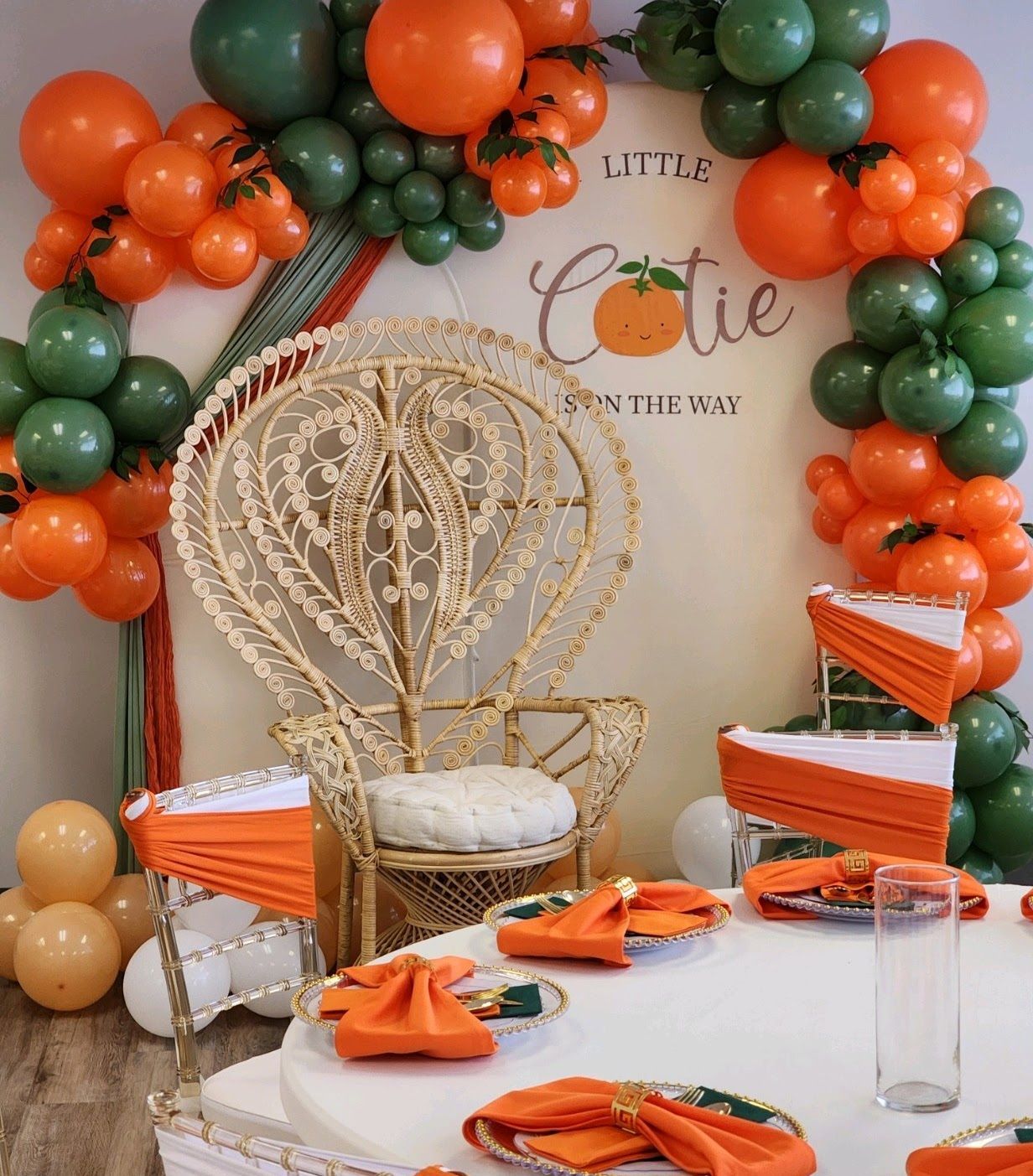 Orange and green balloon arch over a wicker chair and table set for a party with orange and green accents.