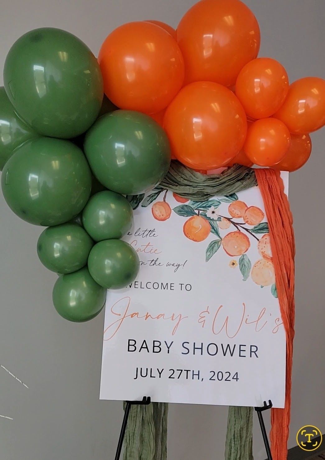 Baby shower sign with orange and green balloon arch, and orange fabric accents.