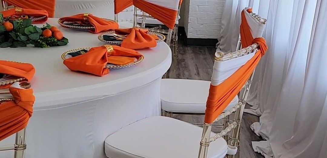 A round white table set for dining, with orange napkins and chair decor, and white chair cushions.