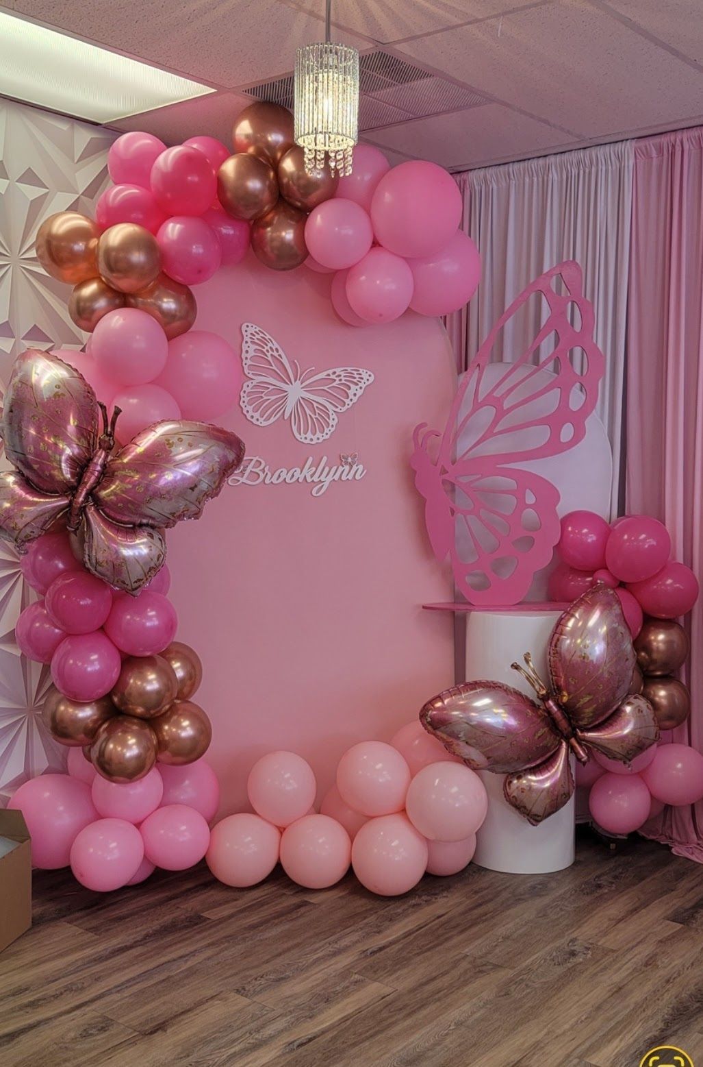 Pink and gold butterfly-themed party backdrop with balloons, cutouts, and decorations.