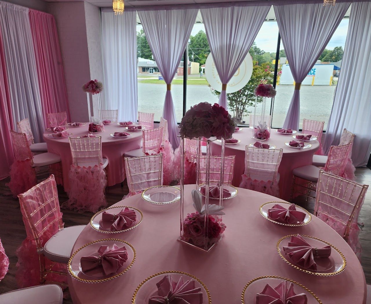 Pink-themed event: round tables with centerpieces, chairs, place settings, and window with curtains.