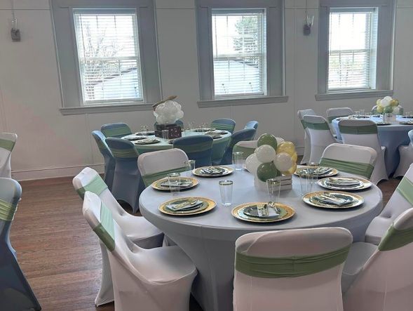 Round tables set for a party in a bright room with windows. White tablecloths, gold plates, and balloon centerpieces.