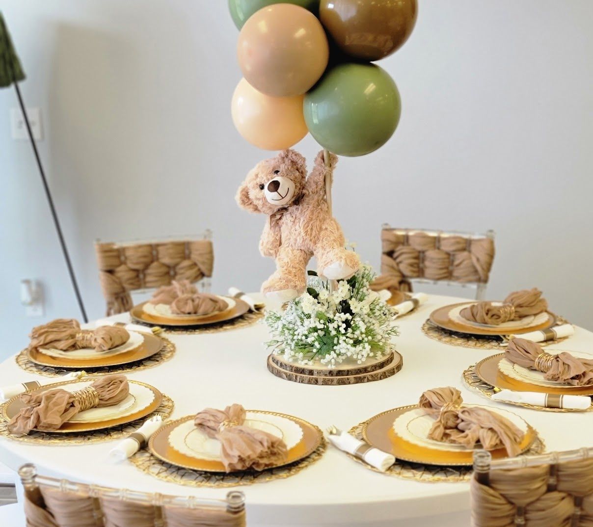 Teddy bear holding balloons atop a party table set with plates, tied napkins, and woven chairs.