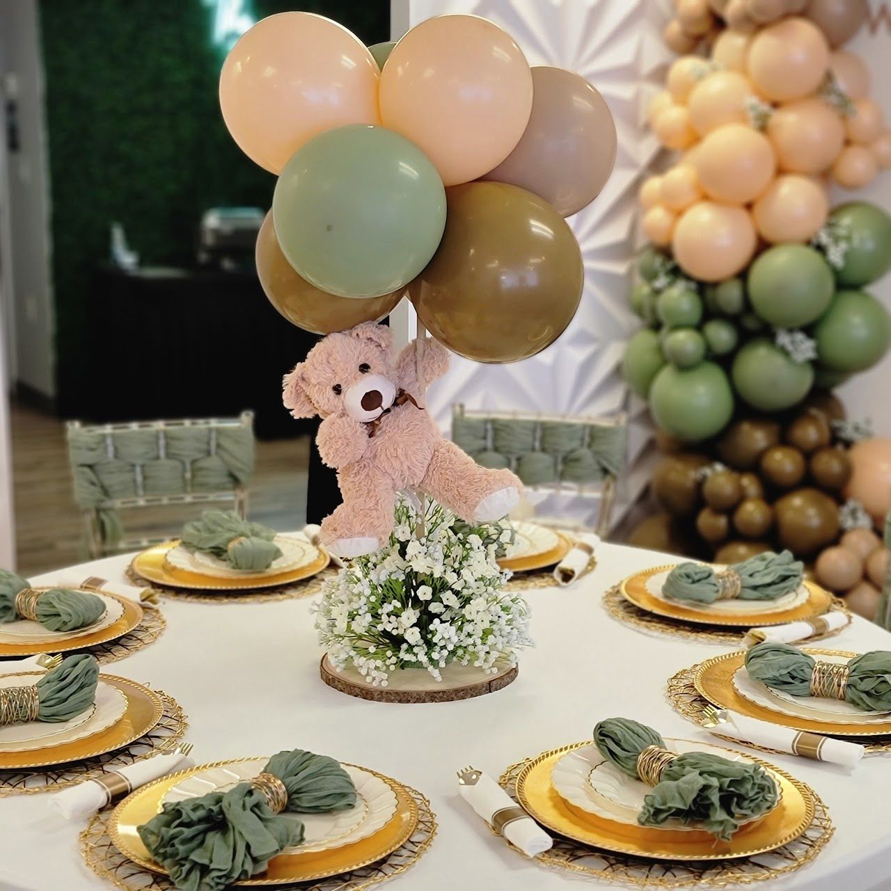 Teddy bear held up by balloons above a table set for a party with gold plates and green napkins.