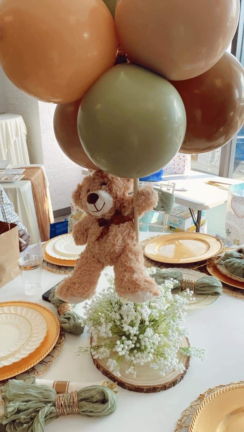 Teddy bear suspended by balloons over a floral arrangement on a table set for a party.