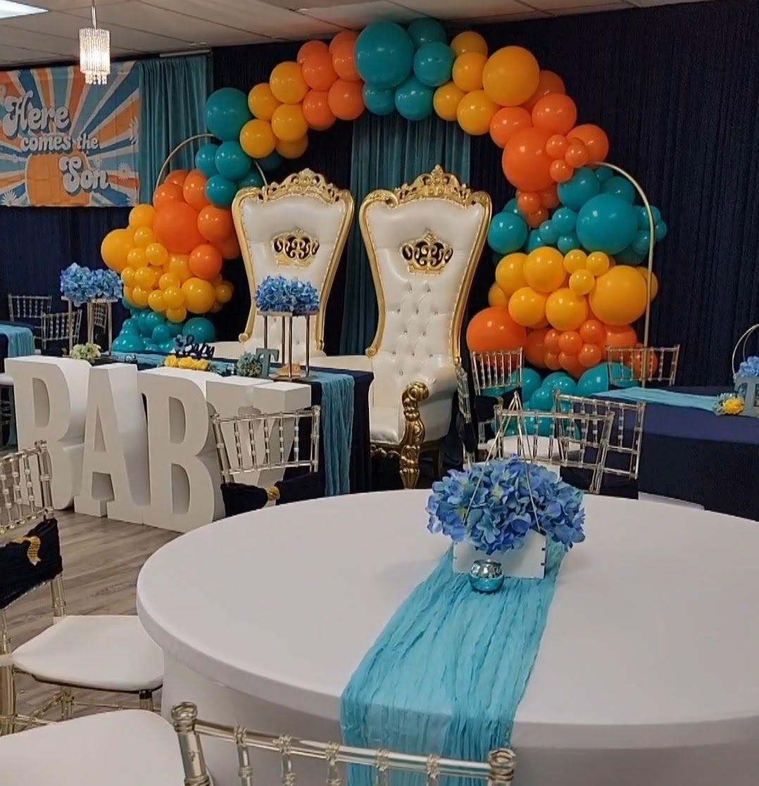 Baby shower with orange, teal, and gold decor: chairs, balloons, table, and floral arrangements.