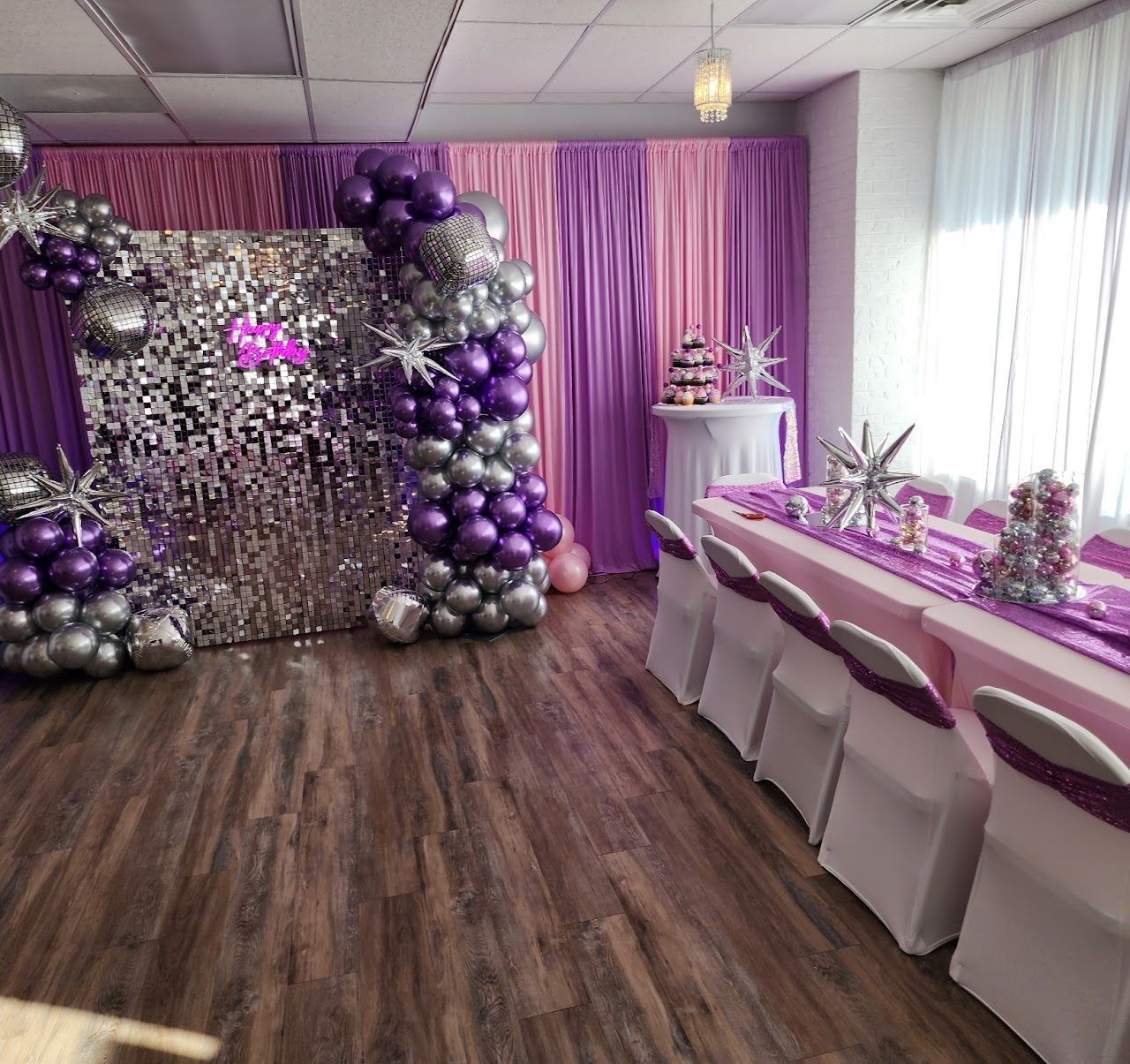 Purple and silver party decorations: balloon arch, sequin backdrop, draped curtains, and decorated tables.
