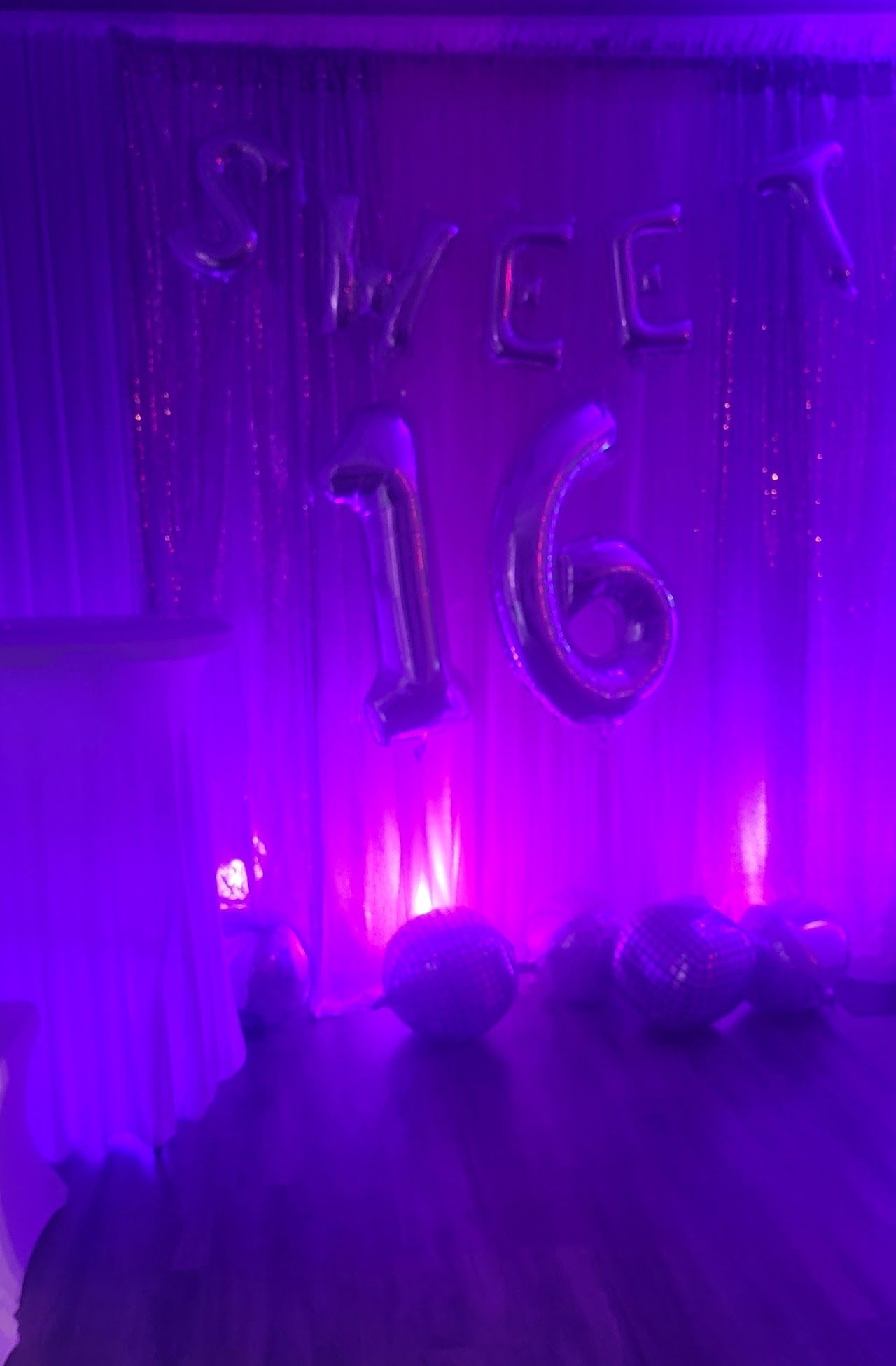 Purple-lit Sweet 16 party decor: foil balloon letters and numbers against a curtain backdrop.