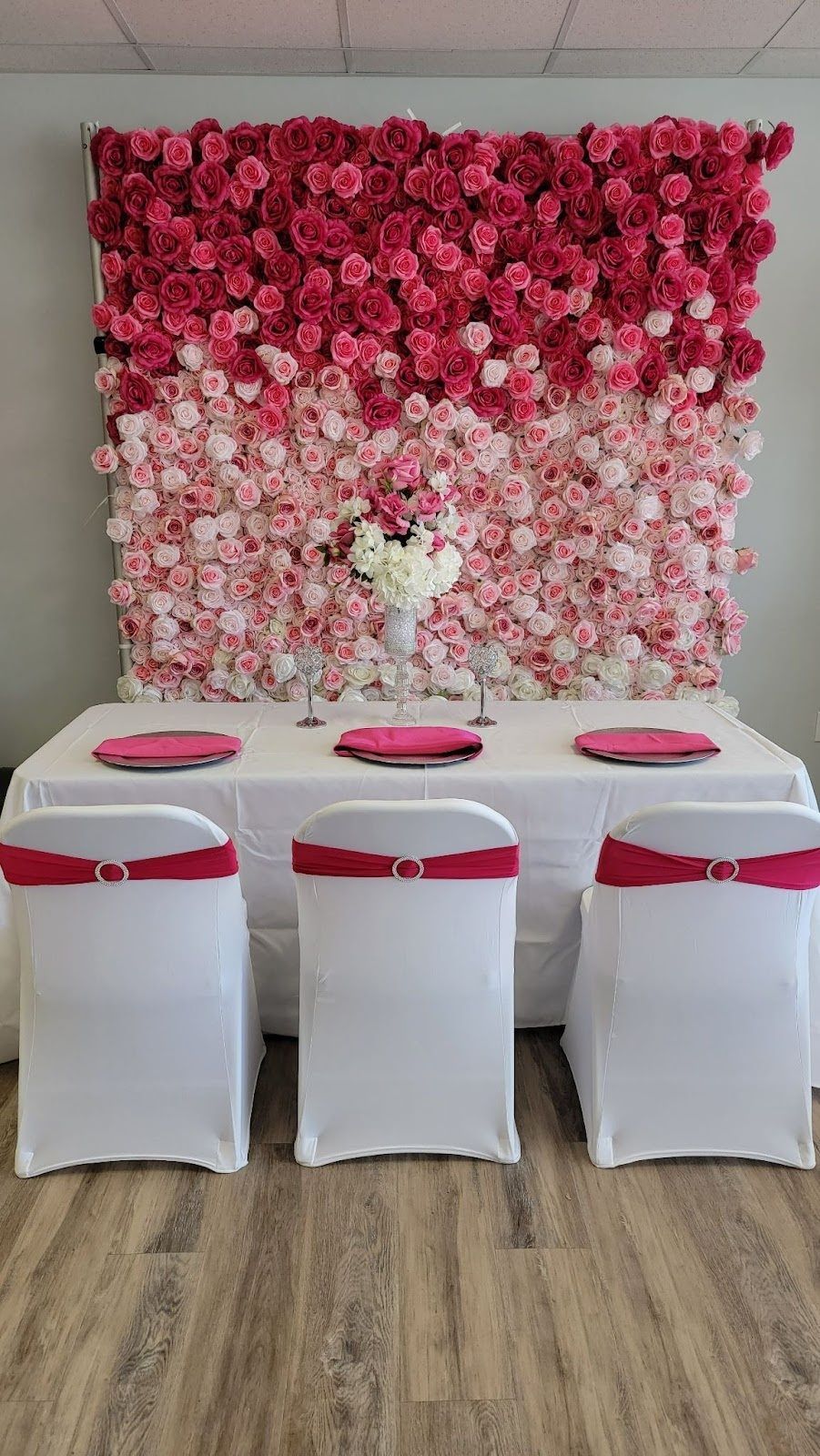 Floral backdrop with table set for three, white chairs with pink sashes, pink and white ombre flowers.