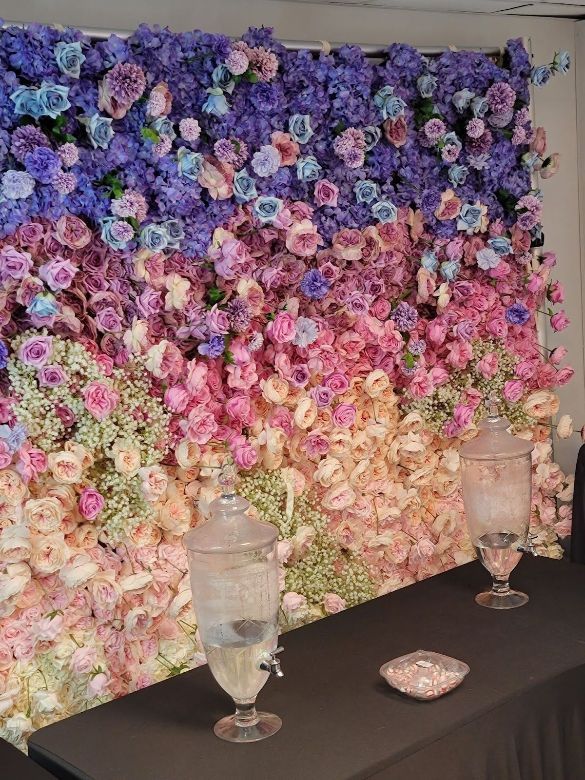 Flower wall backdrop with drink dispensers on a black table.