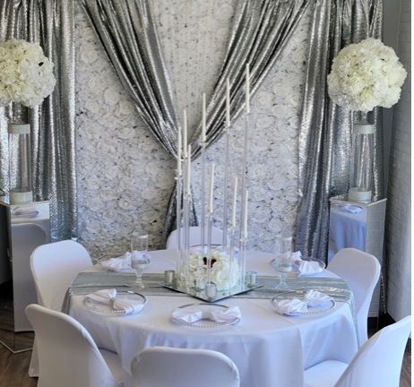 Elegant white and silver event setup: round table with floral centerpiece, white chairs, backdrop with curtains, candles, and pedestals.
