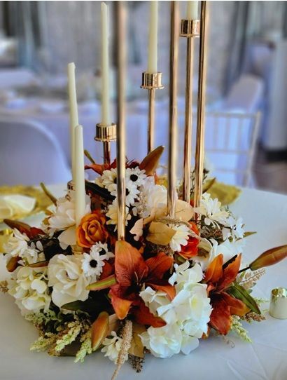 Floral centerpiece with candles on a table. Flowers are white and orange, with gold accents.