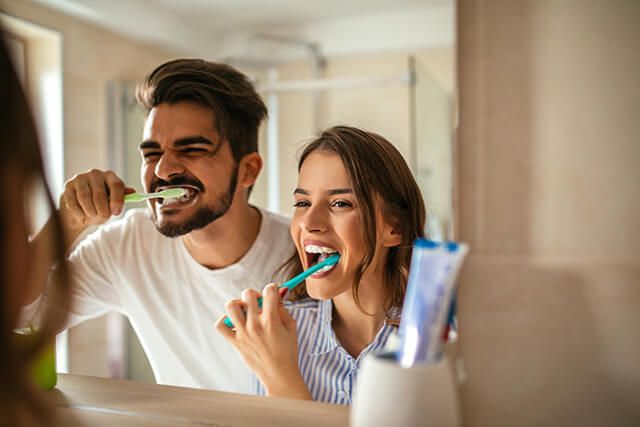 A man and a woman are brushing their teeth in front of a mirror.