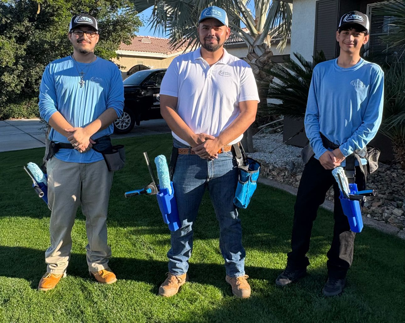 Three people standing on grass holding blue inflatable toys, outdoors in front of a house and car.