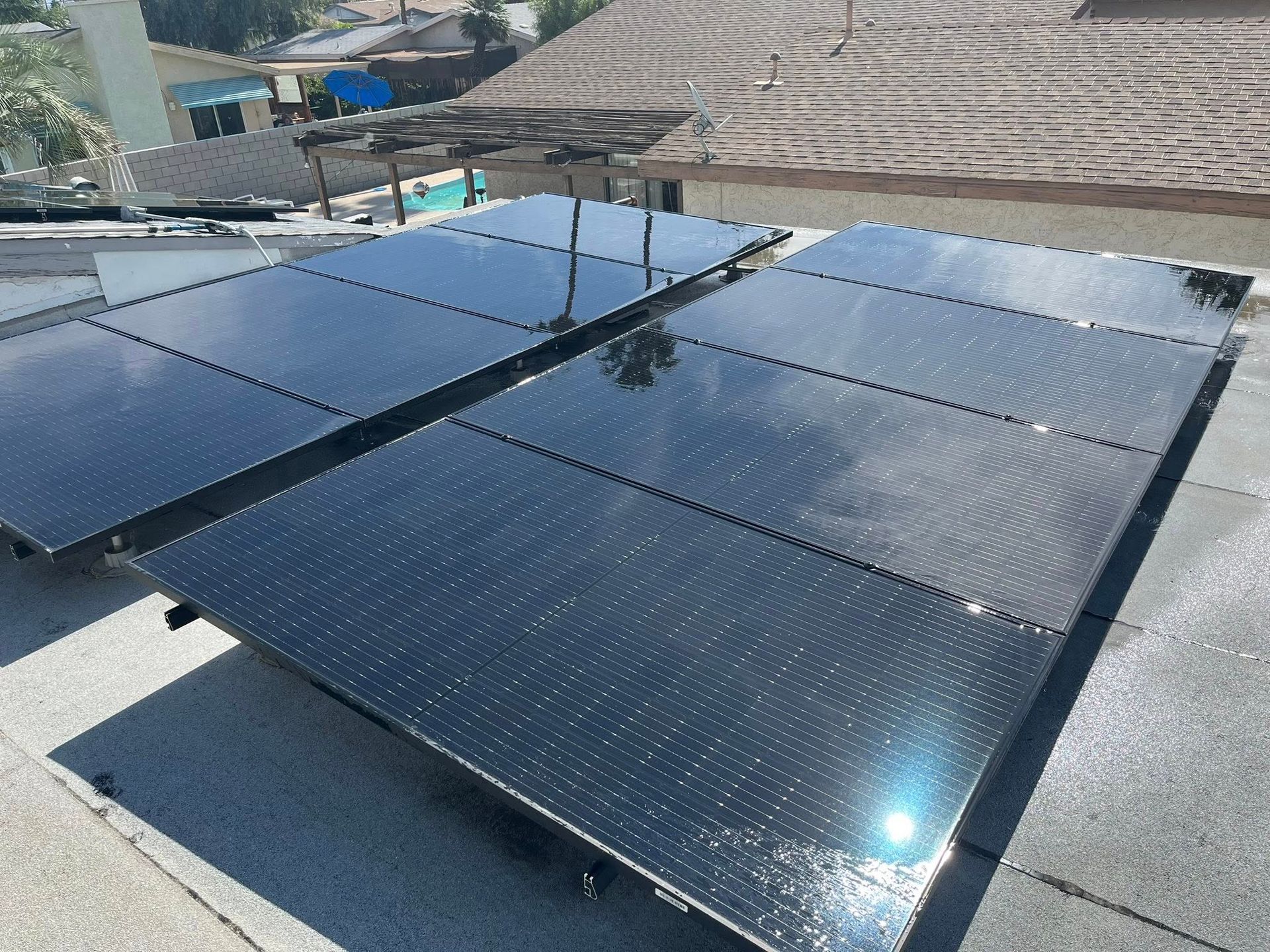 Rows of dark solar panels on a rooftop under bright sunlight, with nearby buildings in the background.