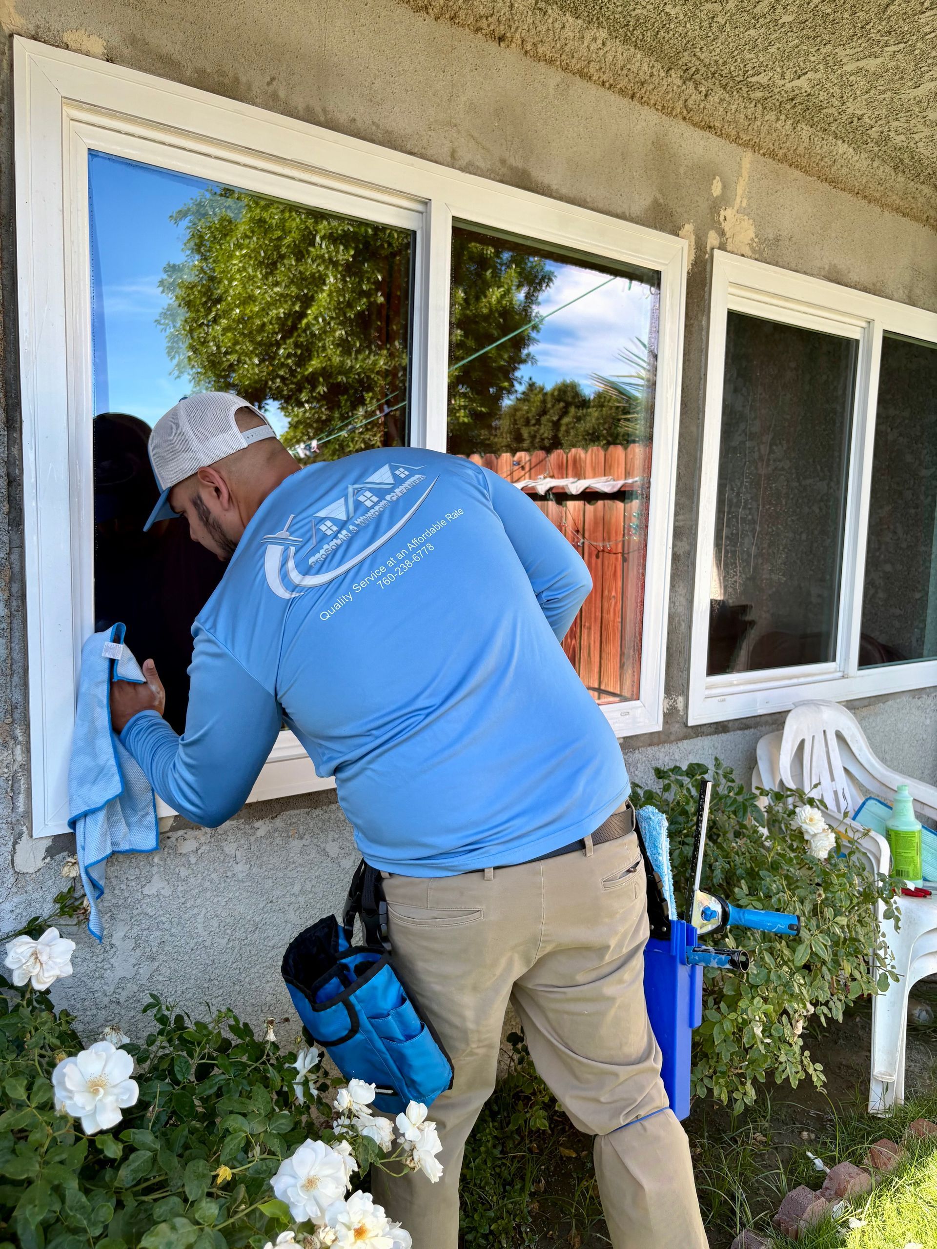 Worker in blue shirt painting an exterior window frame beside a white house and flowers