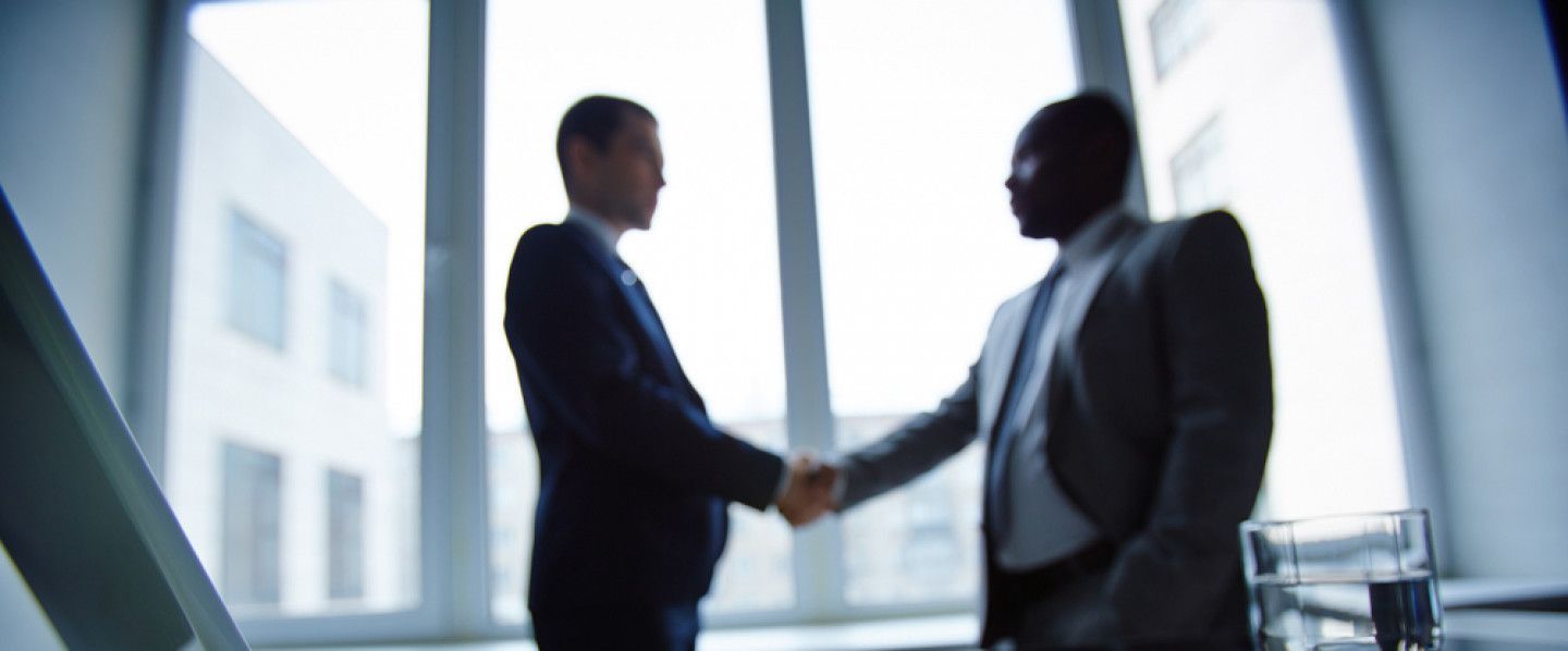 Two men in suits are shaking hands in front of a window.